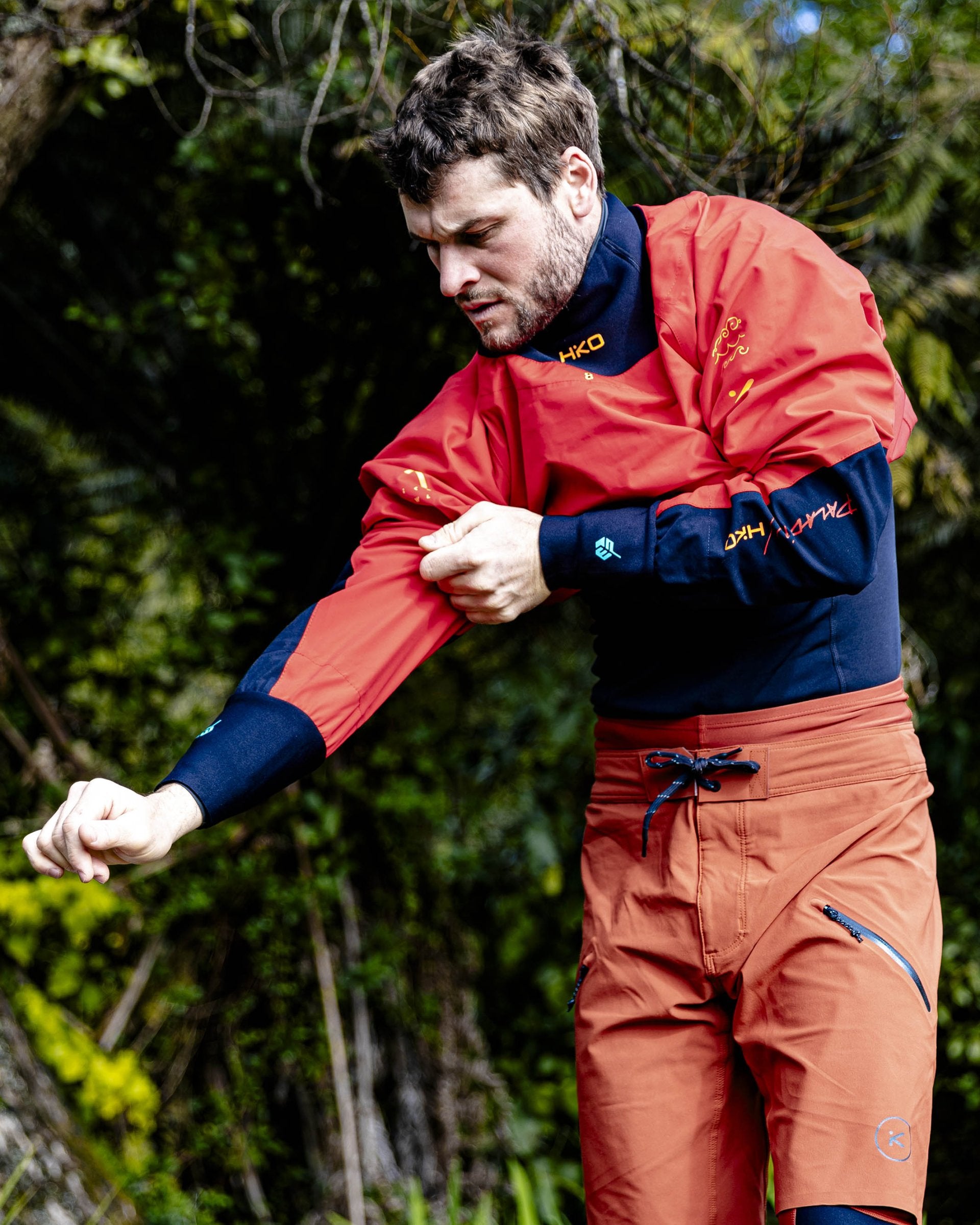 Man wearing a red and blue jacket with a logo, standing outdoors with greenery in the background